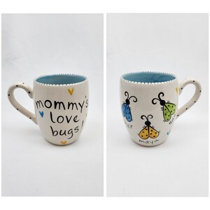 May include: White ceramic mug with a blue rim and a black handle. The mug has a blue heart and yellow hearts painted on it. The text "mommy's love bugs!" is written in black on the mug. The other side of the mug has three ladybugs painted on it, each with a different color and a name written underneath.
