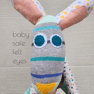May include: A handmade baby safe toy with a gray and blue striped body, pink ears with white and yellow polka dots, and teal felt eyes. The toy is made from fabric scraps and has a yellow felt nose.