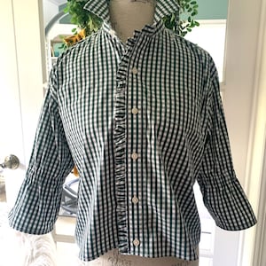 May include: A dark green and white gingham button-down shirt featuring a high collar, three-quarter length sleeves, and a vertical ruffled trim detail running along the front button placket. The shirt is displayed on a mannequin torso.