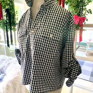 May include: A black and white gingham shirt with a ruffled collar and cuffs. The shirt has two chest pockets and button closures. The sleeves are long and gathered at the cuffs. The shirt is displayed on a mannequin.