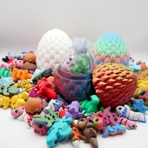 Könnte beinhalten: Eine Sammlung kleiner, bunter, aus Plastik gefertigter Drachenfiguren, einige in eiförmigen Behältern. Die Eier sind mit verschiedenen Mustern verziert, darunter Schuppen, Streifen und Punkte. Im Hintergrund befinden sich auch zwei größere, weiße und pastellfarbene Eier.