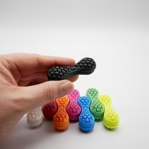 Puede incluir: Un conjunto de diez juguetes de colores, texturados y con forma de cacahuete. Los juguetes están hechos de un material suave y flexible, perfecto para el juego sensorial. Los juguetes están dispuestos en semicírculo, con el juguete negro en el centro.