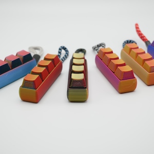 Könnte beinhalten: Fünf bunte Schlüsselanhänger mit jeweils vier Miniatur-Tastaturtasten. Die Schlüsselanhänger sind aus Kunststoff und haben einen Farbverlauf. Die Tasten sind gelb, rot und blau.