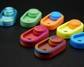 May include: Seven colorful, oval-shaped, 3D printed objects with a raised button on top. The objects are arranged in a row on a black surface. The colors are blue, green, orange, yellow, red, brown, and blue.