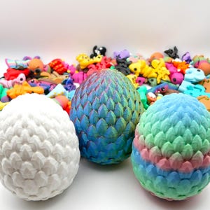 Puede incluir: Tres adornos con forma de huevo de dragón, uno blanco, uno azul iridiscente y otro con un degradado rosa, azul y verde. Los adornos están hechos de un material texturizado y están sobre una superficie blanca.
