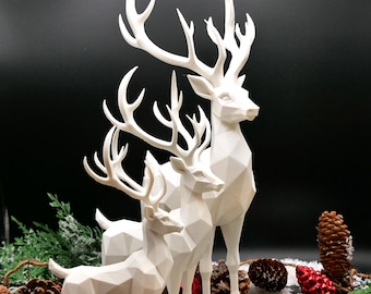 May include: A group of three white geometric deer sculptures with large antlers. The deer are arranged in a cluster, with the largest in the back and the smallest in the front. The sculptures are set against a black background with pinecones and greenery.