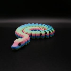 Pode incluir: Um brinquedo de cobra de plástico colorido e segmentado em posição enrolada. A cobra é composta por múltiplas seções, cada uma de uma cor diferente, criando um efeito arco-íris.