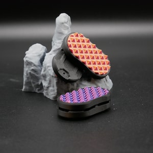 May include: Three small, black, rectangular objects with textured, colorful tops. One has a pattern of orange and red triangles, the other a pattern of purple and blue chevrons. They are displayed near a gray, rock-like sculpture.