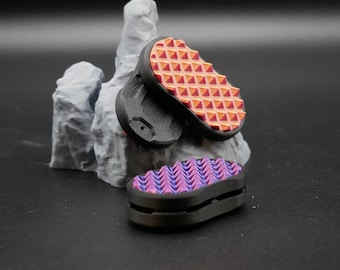 May include: Three small, black, rectangular objects with textured, colorful tops. One has a pattern of orange and red triangles, the other a pattern of purple and blue chevrons. They are displayed near a gray, rock-like sculpture.