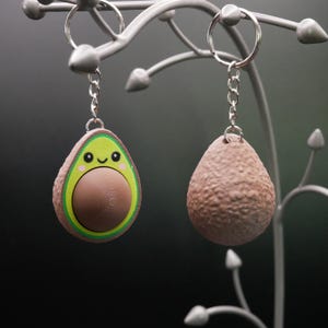 Könnte beinhalten: Zwei avocadoförmige Schlüsselanhänger. Der eine Schlüsselanhänger ist eine komplette Avocado mit einem braunen Kern und einer grünen und gelben Schale. Der andere Schlüsselanhänger ist ein brauner Avocadokern. Beide Schlüsselanhänger sind an silbernen Schlüsselringen befestigt.