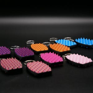 Può includere: Una collezione di portachiavi colorati con un design strutturato e appuntito. I portachiavi sono in vari colori, tra cui rosa, arancione, blu e viola, ciascuno con un portachiavi argentato. Lo sfondo è nero.