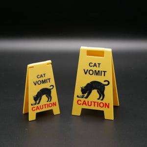 Op de afbeelding: Twee gele, opvouwbare waarschuwingsborden. De borden lezen "CAT VOMIT" en "CAUTION" in zwart en rood. Elk bord heeft een zwart katten silhouet. De borden zijn ontworpen om te waarschuwen voor mogelijke gevaren.