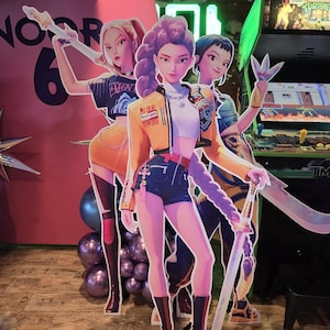 Può includere: Sagome di cartone di tre figure femminili in stile anime, ognuna con un'arma. Le figure sono vestite con abiti alla moda dai colori vivaci. Lo sfondo include uno sfondo rosa, palloncini e un cabinato arcade.