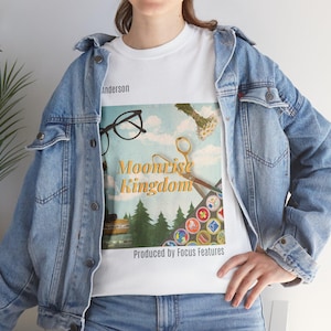 May include: White t-shirt with a graphic design featuring a blue sky, green trees, a pair of glasses, a pair of scissors, and a collection of colorful badges. The text "Moonrise Kingdom" is in yellow and the text "Produced by Focus Features" is in black.