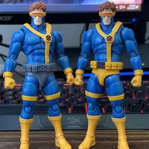 May include: Two action figures of the character Cyclops from the X-Men. The figures are in a dynamic pose, wearing blue and yellow costumes with red visors. The figures have detailed musculature and are standing in front of a computer.