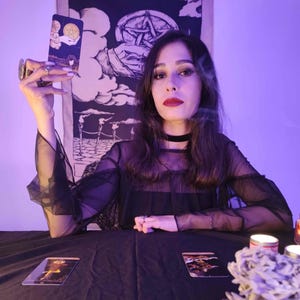 May include: A person holding a tarot card with a gold pentacle design. The person is wearing a black sheer top and a black choker. Two tarot cards are on a black table. A decorative tapestry with a pentagram is in the background.