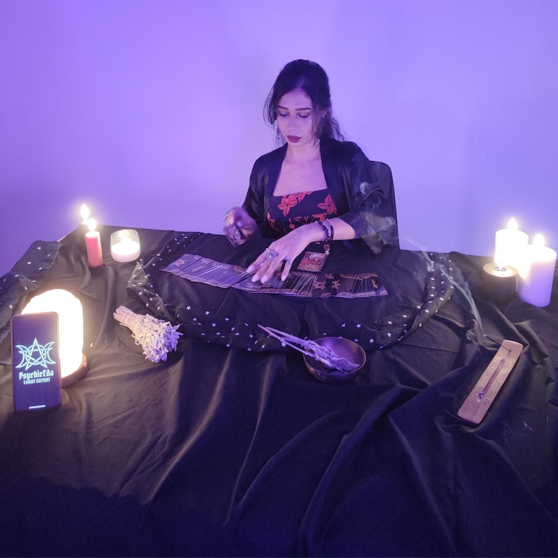 May include: A woman seated at a table with tarot cards, lit by candles and a glowing lamp. The table is draped in black fabric, with a bundle of sage, a bowl, and an incense holder. A smartphone displays the text "Psychic Tarot Expert".