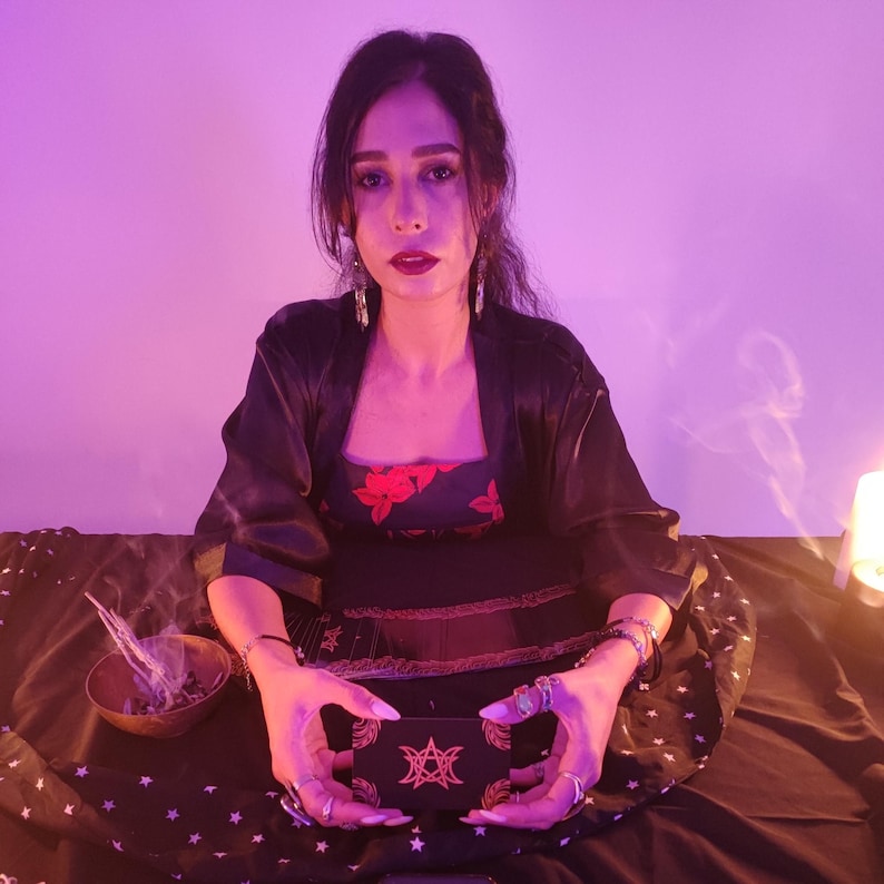 May include: A woman in a black robe and dress holds a black box with a star design. The dress has red floral accents. A small wooden bowl with herbs and incense is nearby. The background is lit with purple and yellow light.