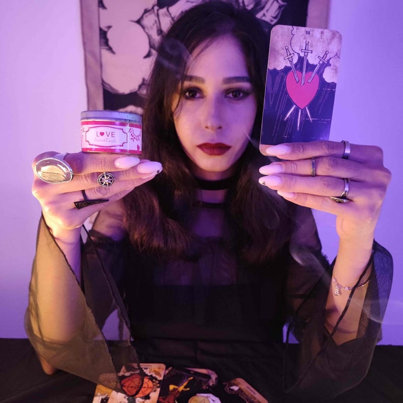 May include: A person holding a tarot card depicting a heart pierced by three swords and a small candle with the word "LOVE". The person is wearing a black top and several rings.