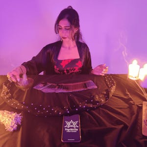 Op de afbeelding: Een persoon in een zwarte jurk en een rood bloemen top zit aan een tafel met tarotkaarten, een kom en een bundel salie. Een smartphone met de tekst "Psychicda Tarot Expert" ligt op de tafel. De sc&egrave;ne wordt verlicht door kaarsen en paars licht.