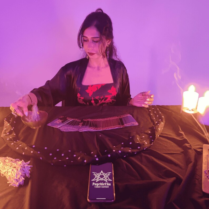 Op de afbeelding: Een persoon in een zwarte jurk en een rood bloemen top zit aan een tafel met tarotkaarten, een kom en een bundel salie. Een smartphone met de tekst "Psychicda Tarot Expert" ligt op de tafel. De sc&egrave;ne wordt verlicht door kaarsen en paars licht.