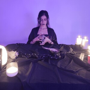 Op de afbeelding: Een persoon houdt tarotkaarten vast, omringd door kaarsen en mystieke voorwerpen. De sc&egrave;ne speelt zich af op een donkere doek met sterrenpatronen, wat een spirituele sfeer cre&euml;ert. De verlichting is zacht paars.