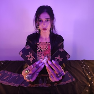 May include: A person holding tarot cards, one featuring a sun illustration. The cards have black backgrounds with gold star and geometric designs. The person wears rings and a black outfit, against a purple backdrop.