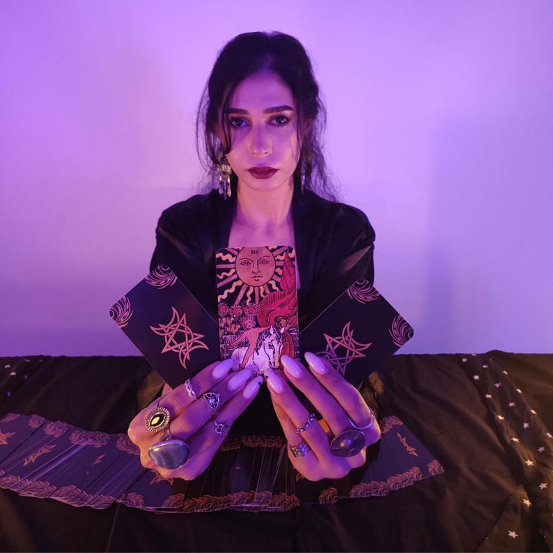 May include: A person holding tarot cards, one featuring a sun illustration. The cards have black backgrounds with gold star and geometric designs. The person wears rings and a black outfit, against a purple backdrop.