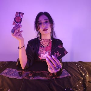 May include: A person holding tarot cards, including one with a wheel design and another with a sun illustration. The cards are displayed against a dark background, with other cards visible. The scene is illuminated with purple lighting.