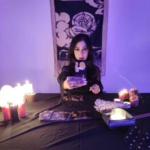 May include: A woman holding a tarot card, with a black tablecloth and candles. A tapestry with a pentagram design hangs in the background. The scene is illuminated by candlelight, creating a mystical atmosphere. Tarot cards are laid out on the table.