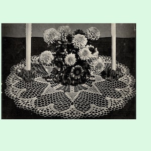 May include: A white crocheted doily with a delicate floral pattern, featuring a centrepiece of white flowers and two candlesticks.