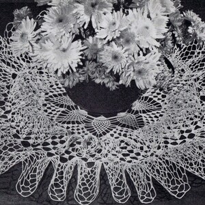 May include: A white crocheted doily with a scalloped edge, featuring a delicate floral design. The doily is displayed on a dark background with white flowers.