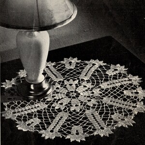 May include: A white crocheted doily with a delicate floral design sits on a dark surface under a lamp.