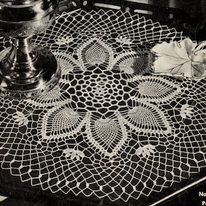 May include: A white crocheted doily with a delicate floral design. The doily is approximately 12 inches in diameter and features a central motif of six overlapping petals. The doily is displayed on a black surface with a silver pitcher and a white leaf-shaped dish.