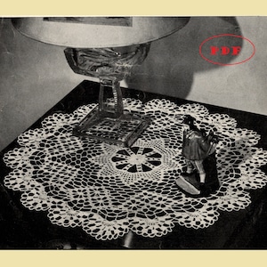 May include: A white crocheted doily with a delicate floral pattern sits on a dark surface. A glass lamp base and a small figurine are also visible.