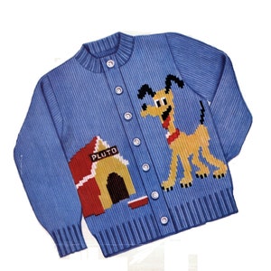 Op de afbeelding: Een blauwe gebreide trui met een cartoon afbeelding van Pluto de hond en zijn hondenhok. Het hondenhok heeft een rood dak en de tekst "Pluto" op de voorkant.