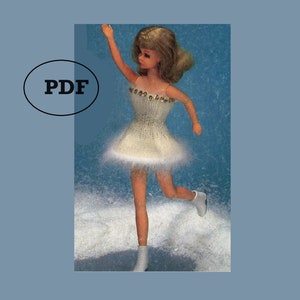 May include: A white knitted ice skating dress with a beaded bodice for a doll. The doll is wearing white skates and is posed in a skating position.