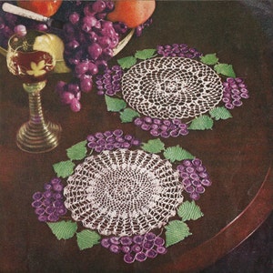 May include: Two white crocheted doilies with a grapevine design. The doilies are on a dark brown wooden table with a bowl of grapes and a glass of wine.