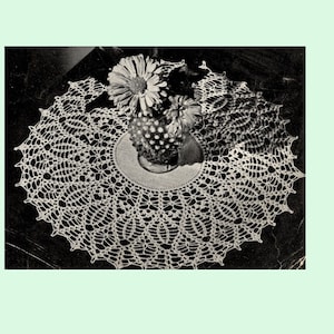 May include: A white crocheted doily with a delicate floral pattern. The doily is placed on a dark surface with a vase of flowers in the centre.