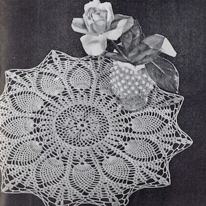 May include: A white crocheted doily with a delicate, intricate design. The doily is circular and features a central motif with radiating spokes. A white rose in a clear glass vase sits on top of the doily.