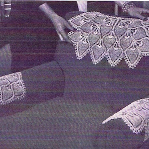 Könnte beinhalten: Ein cremefarbener Spitzenkragen und passende Manschetten. Der Kragen hat einen gewellten Rand und ein sich wiederholendes blattartiges Muster. Die Manschetten haben ein ähnliches Design. Die Spitze scheint handgefertigt zu sein, auf dunkler Oberfläche.