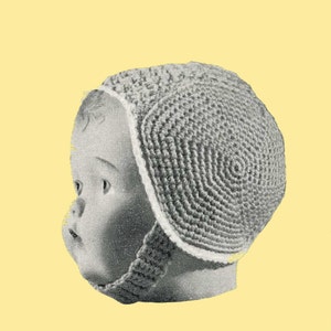 Può includere: Un berretto per neonato in crochet grigio con le alette per le orecchie. Il berretto è indossato sulla testa di una bambola.