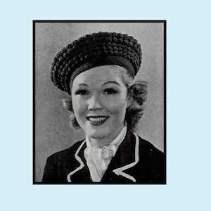 May include: A black beret with a textured surface. The beret is worn by a person with short, wavy hair. The person is wearing a dark jacket with a white collar and white trim.