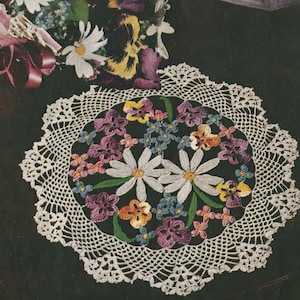 Pode incluir: Um tapete de crochê branco com um desenho floral que apresenta margaridas, violetas e miosotis em tons de roxo, azul, amarelo e verde.