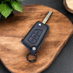 Può includere: Un portachiavi in ​​pelle nera con un anello portachiavi argentato e tre pulsanti etichettati "B", "1" e "D". Il portachiavi è su una superficie di legno.