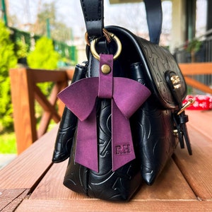 May include: A black leather handbag with an embossed pattern and a top handle, featuring a dark purple leather bow charm attached to a gold-toned metal ring. The bow has the letters RH embossed on one of its hanging tails and a brass rivet.