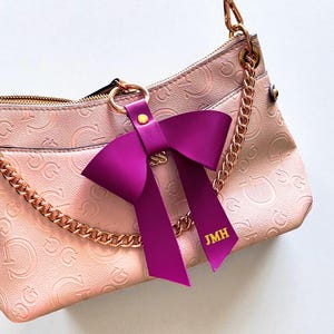 May include: A light pink handbag with a repeating embossed G logo pattern, featuring a gold-tone chain strap and a large, deep purple leather bow charm attached to a gold ring. The bow has the gold-stamped initials JMH on one of its tails.