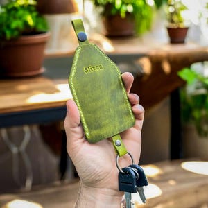 May include: A green leather key holder with the word "Sliven" embossed on the front. The key holder has a metal ring attached to the bottom for keys.