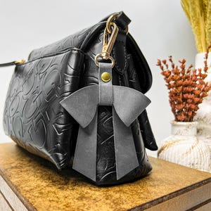 May include: A black handbag with a repeating embossed pattern and a gold-tone metal clasp. Attached to the clasp is a grey leather bow charm featuring a small gold-tone rivet. The handbag has a structured shape with a front pocket and a flap closure.