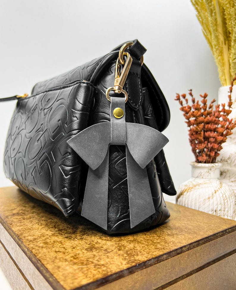 May include: A black handbag with a repeating embossed pattern and a gold-tone metal clasp. Attached to the clasp is a grey leather bow charm featuring a small gold-tone rivet. The handbag has a structured shape with a front pocket and a flap closure.